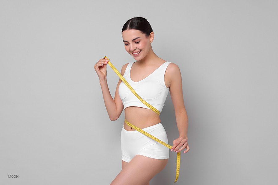 a woman in white underwear smiles as she wraps a measuring tape around her body